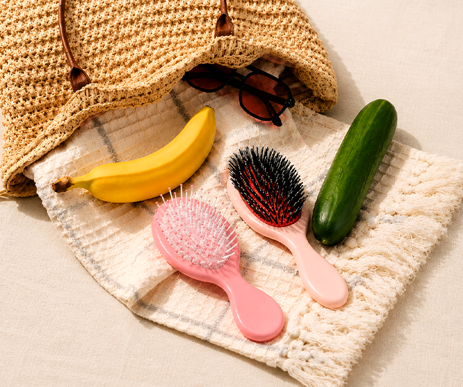 Pink hairbrushes, banana, sunglasses, and cucumber on a textured surface with a woven bag.
